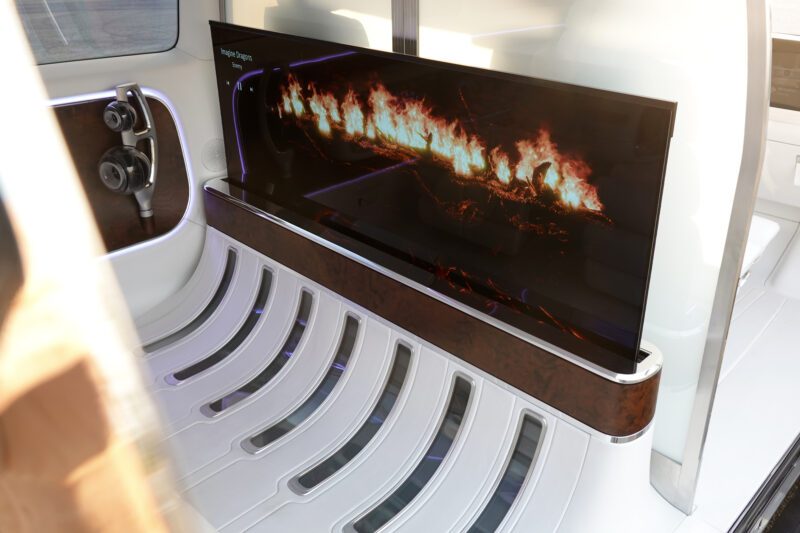 A large screen displaying a digital fireplace is mounted inside the Mercedes-Benz Vision V concept luxury van, featuring a modern interior with white and wood accents.