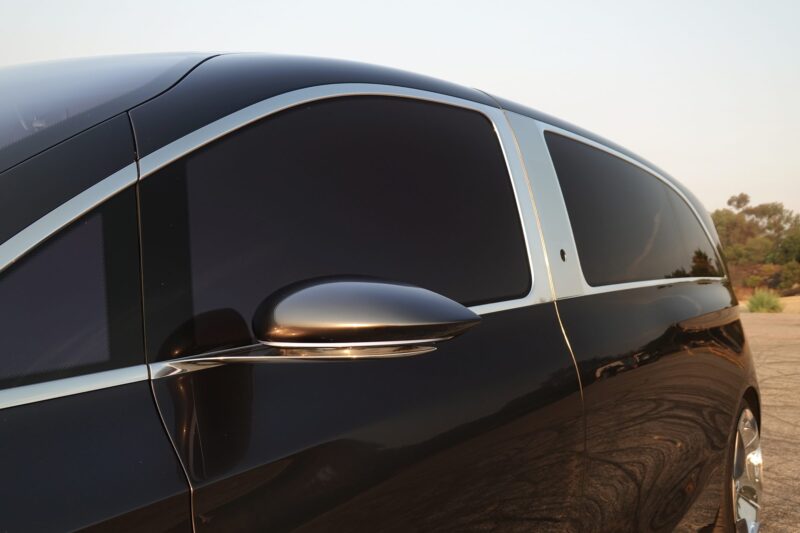 Close-up of the side of a black Mercedes-Benz Vision V concept van with tinted windows, chrome trim, and a side mirror, parked outdoors on a clear day, showcasing modern van luxury.