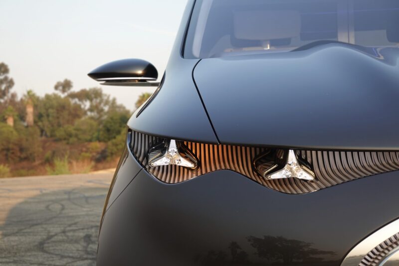 Close-up of a modern black car's front end, inspired by the sleek lines of the Mercedes-Benz Vision V concept, with two angular headlights and a curved hood. Trees and sky appear blurred in the background.