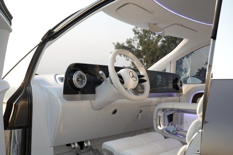 Interior of the Mercedes-Benz Vision V concept van showcases luxury with a white dashboard, steering wheel, digital displays, and ambient lighting—all visible through the open driver's door.
