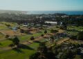 Aerial view of a golf course with scattered trees, parked cars, tents, and nearby buildings; coastline and ocean visible in the background during Mecum Auctions at Monterey Car Week.
