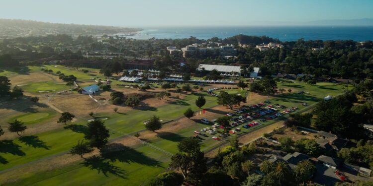 Aerial view of a golf course with scattered trees, parked cars, tents, and nearby buildings; coastline and ocean visible in the background during Mecum Auctions at Monterey Car Week.