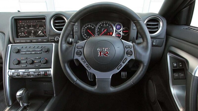 Close-up view of a Nissan R35 GT-R car interior, showcasing the steering wheel, dashboard, center console, gear shifter, and control buttons—capturing the spirit of a next generation before the end of an era.