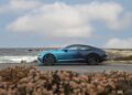 A blue Bentley with a stunning Ombre By Mulliner paint finish is parked on a coastal road, set against the ocean and cloudy sky; plants brighten the foreground.