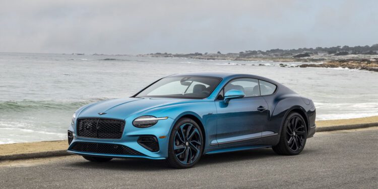 A blue Bentley sports car with an Ombre By Mulliner paint finish is parked on a coastal road, set against the ocean and a cloudy sky in the background.