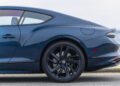 Side view of a dark blue Bentley luxury coupe with an Ombre By Mulliner paint finish, focusing on the rear wheel, door, and window, set against the ocean in the background.