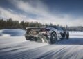 A black and white camouflaged McLaren 750S Le Mans speeds down a snowy road, surrounded by snow-covered trees and a blue sky—perfect for making an entrance at Exotics On Broadway.