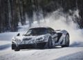 A McLaren 750S Le Mans with camouflage wrap drives at high speed on a snowy road, kicking up snow as it drifts around a corner—an Exotics On Broadway spectacle in winter.