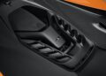 Close-up view of a McLaren 750S Le Mans engine cover with visible vents and the McLaren logo, surrounded by carbon fiber bodywork and an orange exterior section at Exotics On Broadway.