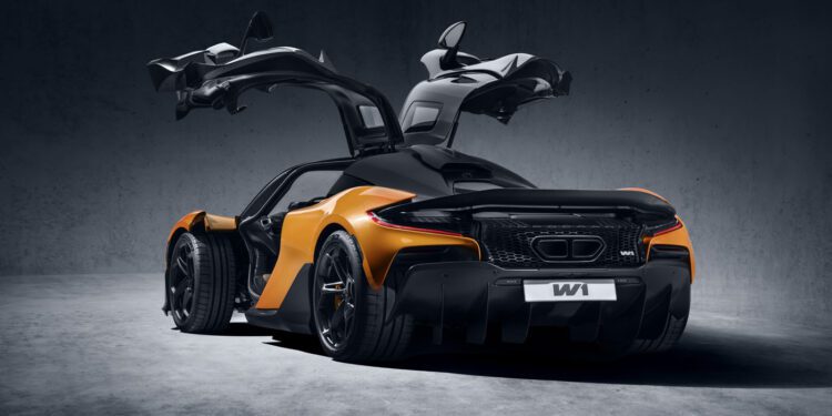 A sleek orange and black McLaren 750S Le Mans sports car with its distinctive butterfly doors open, shown from the rear in a dimly lit studio setting.