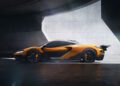 A sleek orange McLaren sports car with black accents is parked indoors against a large, gray concrete wall, illuminated by natural light—reminiscent of the stunning 750S Le Mans edition.