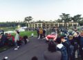 A crowd watches a red classic convertible car driving on a paved path at an outdoor event near a large building and manicured lawns.