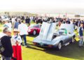 A crowd gathers at a car show on a lawn, with classic cars on display, including a light blue convertible with its hood open in the foreground.
