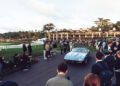 A crowd gathers outdoors near a large building during Car Week to watch a classic convertible Maserati drive along a paved path.
