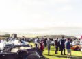 A large crowd gathers outdoors on a grassy field, surrounded by classic cars and a striking Maserati GT2 Stradale, with hills and a cloudy sky in the background.