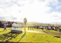 People gather near the Pebble Beach Concours d’Elegance sign during Car Week, with vintage cars—including a Maserati—and a clock on a sunny day.