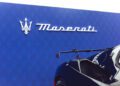 A Maserati logo and brand name are displayed on a blue wall, with part of a black GT2 Stradale sports car visible in the foreground—an eye-catching scene perfect for Car Week enthusiasts.
