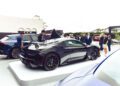 A black Maserati GT2 Stradale on display outdoors, surrounded by people viewing other vehicles during Car Week at an auto show event.