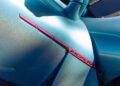 Close-up of a blue McLaren sports car’s body at Car Week, showcasing a striking red trim detail and the McLaren badge near the side mirror.