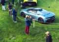People photograph a silver Maserati GT2 Stradale displayed on a grass field, surrounded by a white chain barrier, with another yellow sports car in the background at Car Week.