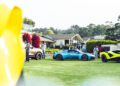 A group of people stand near several luxury cars, including a blue Maserati GT2 Stradale, on a grassy field in front of modern houses and trees during Car Week.