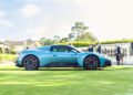 A blue Maserati sports car is parked on a grassy area during Car Week, with people standing nearby and buildings in the background.