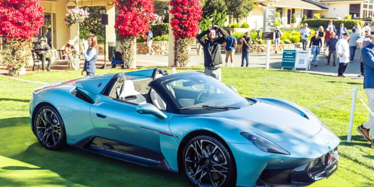 A light blue Maserati GT2 Stradale convertible sports car displayed on a grassy area during Car Week, with people and buildings in the background.
