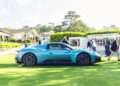 A blue Maserati GT2 Stradale is parked on a green lawn near a building during Car Week, with people standing and walking in the background.