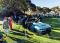 A blue Maserati GT2 Stradale with its doors open is displayed on a grassy area, surrounded by people observing and taking photos at an outdoor car show.