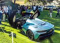 A silver Maserati GT2 Stradale with butterfly doors open is displayed on a grassy lawn among a crowd at Car Week's outdoor show.