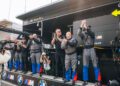 A group of BMW M Motorsport team members in racing uniforms stand on a pit wall, clapping and cheering, with a cameraman recording the scene.
