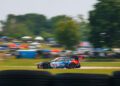 A race car with iHeartRadio branding speeds along a racetrack, with blurred spectators and trees in the background, indicating motion.