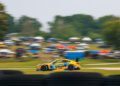 A yellow and blue race car speeds along a track, with blurred spectators and tents in the background, emphasizing motion.