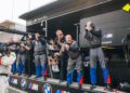 A group of BMW Motorsport team members in uniforms stand on a pit lane, clapping and cheering, as a cameraman films them.
