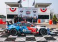 A race car with iHeartRadio branding is parked on a checkered podium in front of a winning race team posing for a group photo at the WeatherTech SportsCar Grand Prix event.