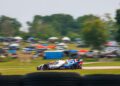 A race car speeds along a track with blurred tires in the foreground and spectators with tents and cars in the background.