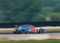 A blue and red race car with iHeartRadio branding speeds along a racetrack, motion blur in the background indicating fast movement.