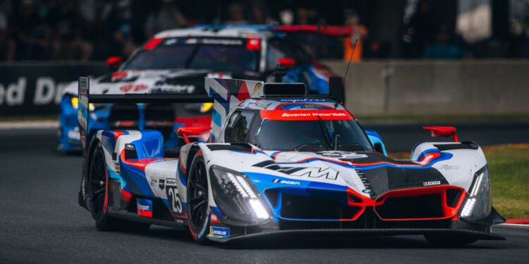 A white, blue, and red race car leads another car on a racetrack during a motorsport event, with spectators and barriers visible in the background.