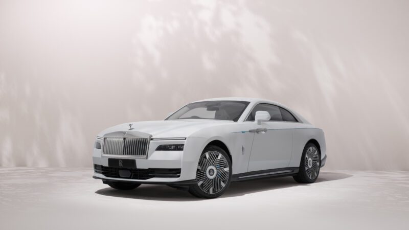 A white Rolls-Royce luxury coupe with a distinctive grille and sleek design is parked in front of a light-colored, softly lit background.
