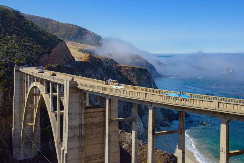 Bixby Creek Bridge spans a coastal canyon along Highway 1 in California, where cars—perhaps even those showcased at Monterey Car Week 2025 by duPont REGISTRY—cross as fog rolls in over cliffs and the Pacific Ocean below.