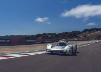 A white Porsche 963 RSP race car drives on a racetrack with hills and trees in the background under a blue sky, capturing the excitement of Car Week.