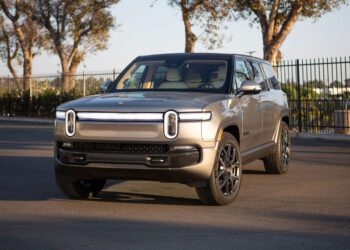 A silver Rivian R1S electric SUV is parked on a paved surface near a black metal fence and trees under daylight, ready to turn heads at Monterey Car Week.