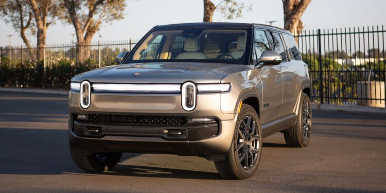 A silver Rivian R1S electric SUV is parked on a paved surface near a black metal fence and trees under daylight, ready to turn heads at Monterey Car Week.
