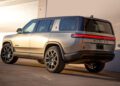 A silver Rivian SUV, shown from the rear three-quarter view, is parked near a light-colored wall in bright sunlight—a striking sight for anyone attending Monterey Car Week or watching an exclusive auction.