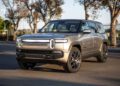 A silver Rivian R1S electric SUV is parked on a paved road near a black metal fence and trees in the background, ready to turn heads at Monterey Car Week or hit the auction block.