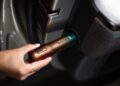A hand removes a metallic flashlight labeled "Rivian" from a built-in charging slot on a car door panel, highlighting an innovative feature showcased at Monterey Car Week.