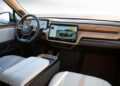 Interior of a Rivian vehicle showing a modern dashboard with two digital screens, wood trim, white seats, and a view of the steering wheel—an innovative design previewed at Monterey Car Week and now available at auction.