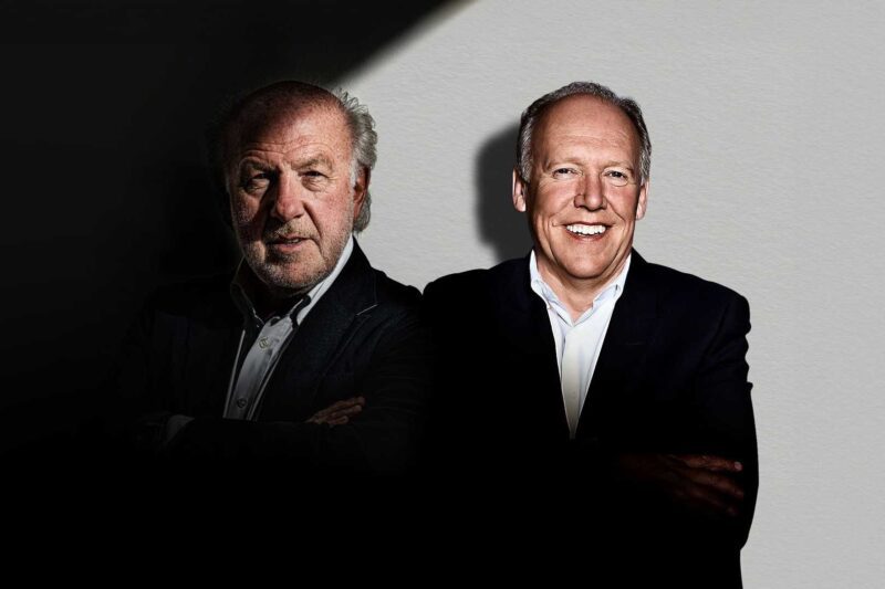 Two men in suits stand side by side against a light-colored wall, one partially in shadow and the other fully illuminated, both with arms crossed—much like the unveiling of the Collector’s Racing Sim: Ex-Jag Designer and Prodrive's limited edition rig.