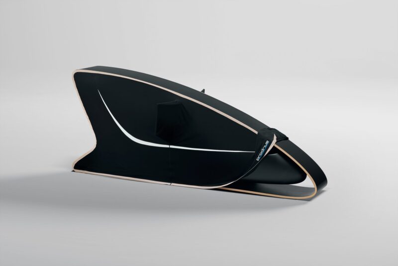 A black, aerodynamic bike travel case with a curved design, wooden trim, and a carrying handle sits on a plain light background—crafted with the same attention to detail as a Collector’s Racing Sim: Ex-Jag Designer and Prodrive Unveil a Limited Edition Rig With B&O Sound.