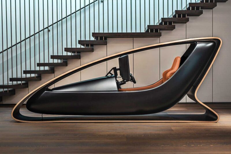 A modern Collector’s Racing Sim setup with a sleek black and wood frame, tan seat, steering wheel, and monitor—this limited edition rig by ex-Jag designer and Prodrive with B&O sound sits perfectly under a wooden staircase.